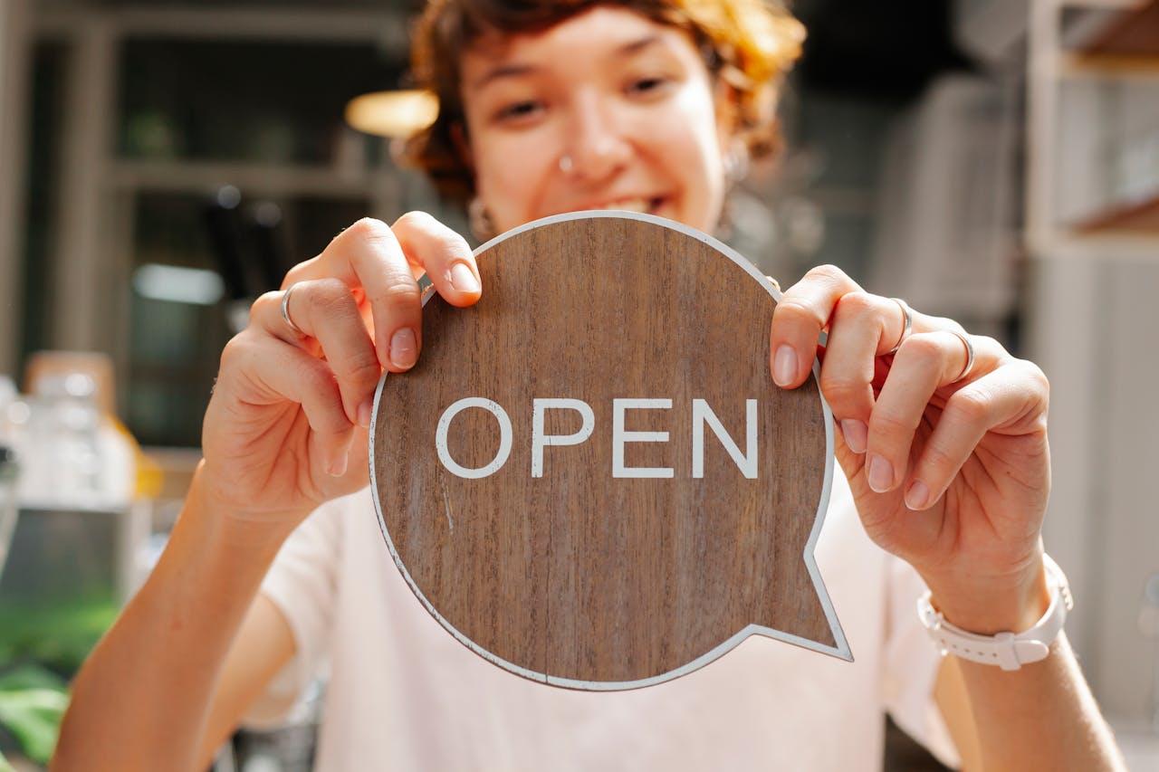 our-experience Happy young woman worker in casual clothes standing in light cafe and showing small sign with word open