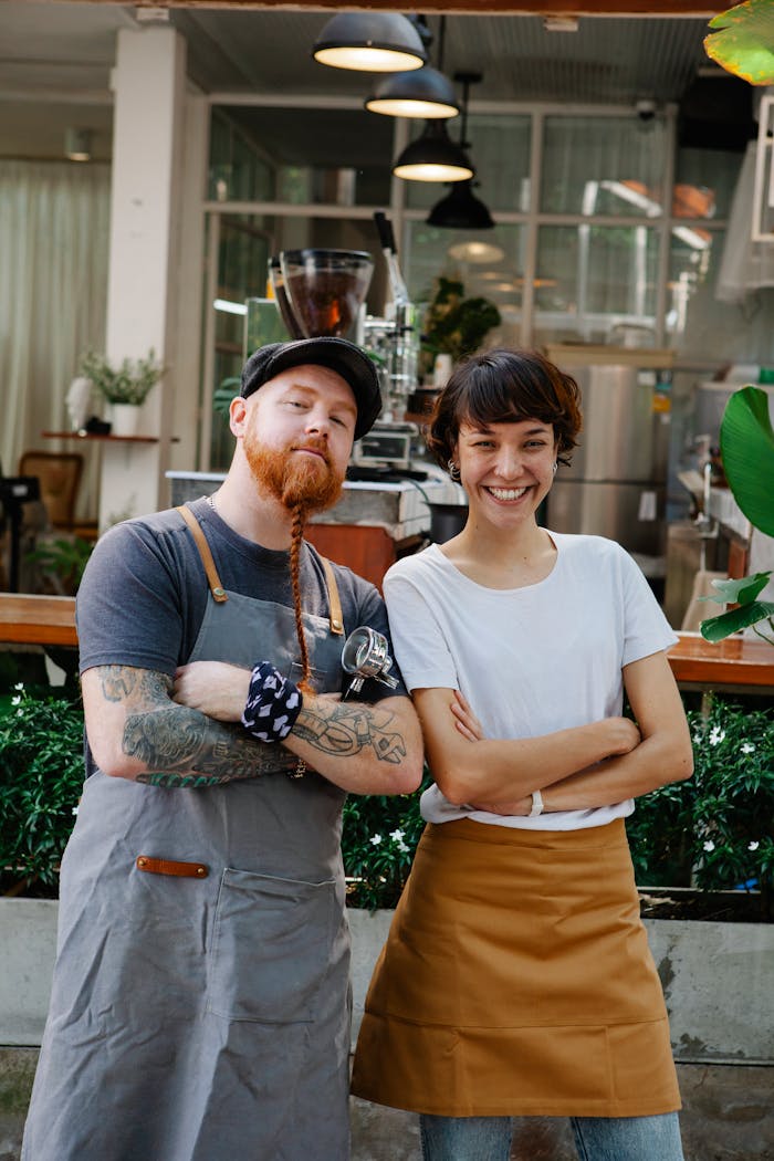 services-03 Couple of confident colleagues in casual clothes and aprons spending time in street near cafe with portafilter near plants while looking at camera in daytime